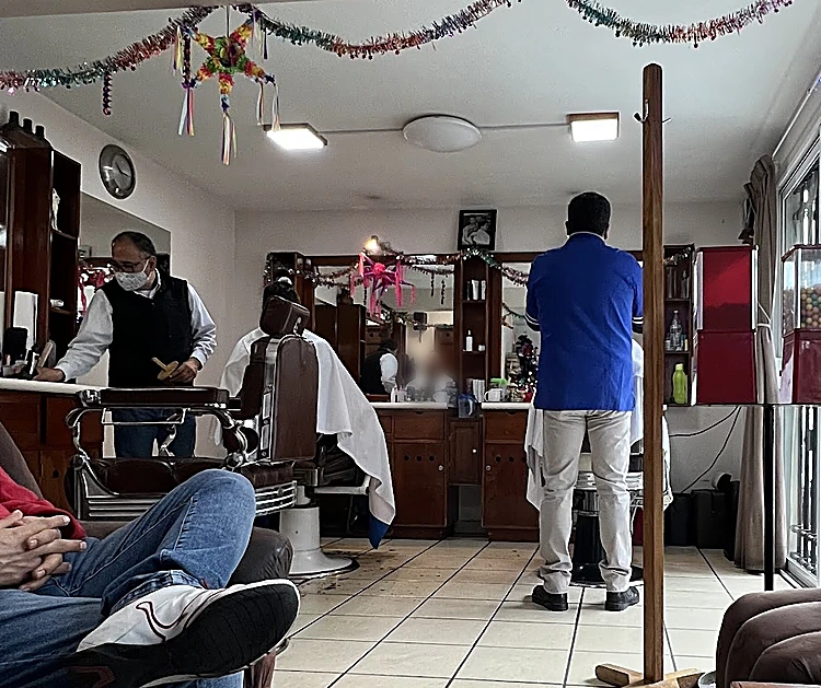 Interior de la barbería: sillón clásico de cuero, tocadores y espejos de madera, piso de loseta y dos barberos atendiendo