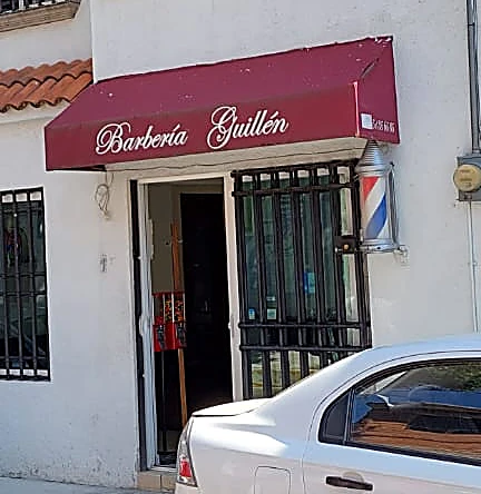 Entrada de la barbería con el toldo guinda que dice Barbería Guillén en letra blanca y el poste de barbero junto a la puerta